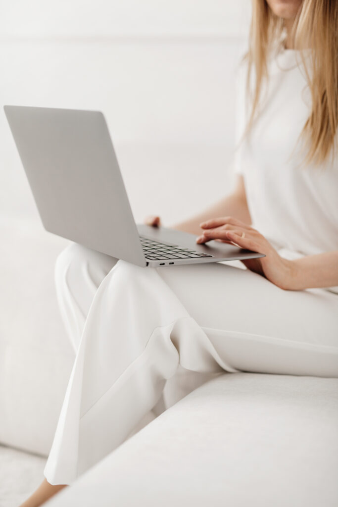 A person sits on a white couch in white clothes, browsing a website platform for wedding planners on their silver laptop. Their face is not fully visible.