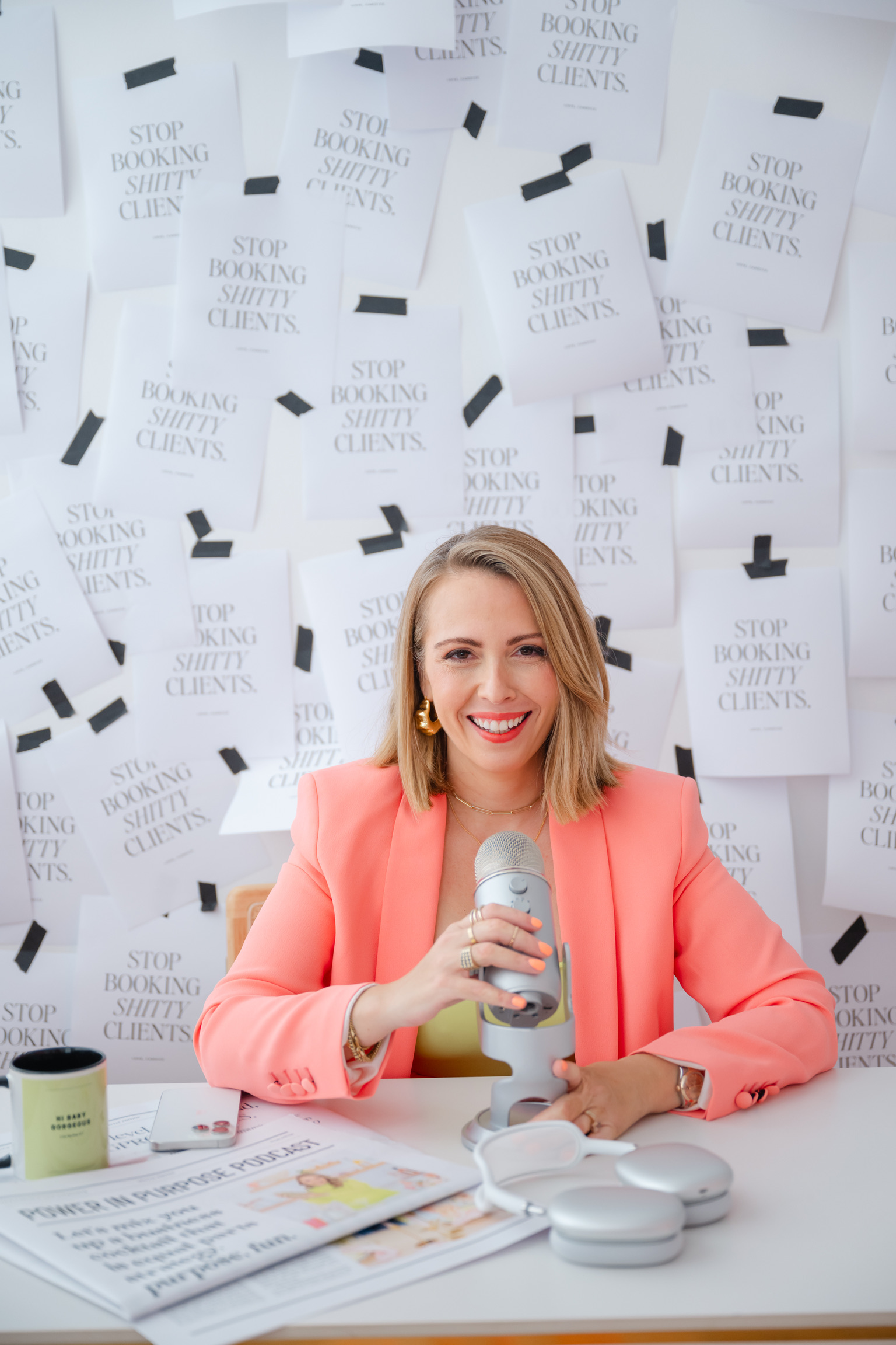 A woman in a pink blazer sits at a desk with a microphone, headphones, and papers—prepping for her brand photoshoot as a wedding planner. Behind her, the wall is covered with taped papers reading “STOP BOOKING SH*TTY CLIENTS.”.