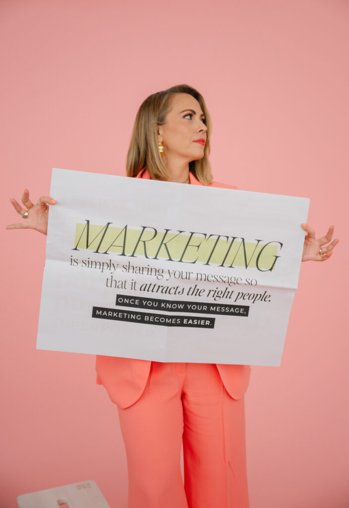 A woman in a coral suit holds a sign with marketing text in front of a pink background, perfect for a brand photoshoot. The sign reads, "Marketing is simply sharing your message so that it attracts the right people.