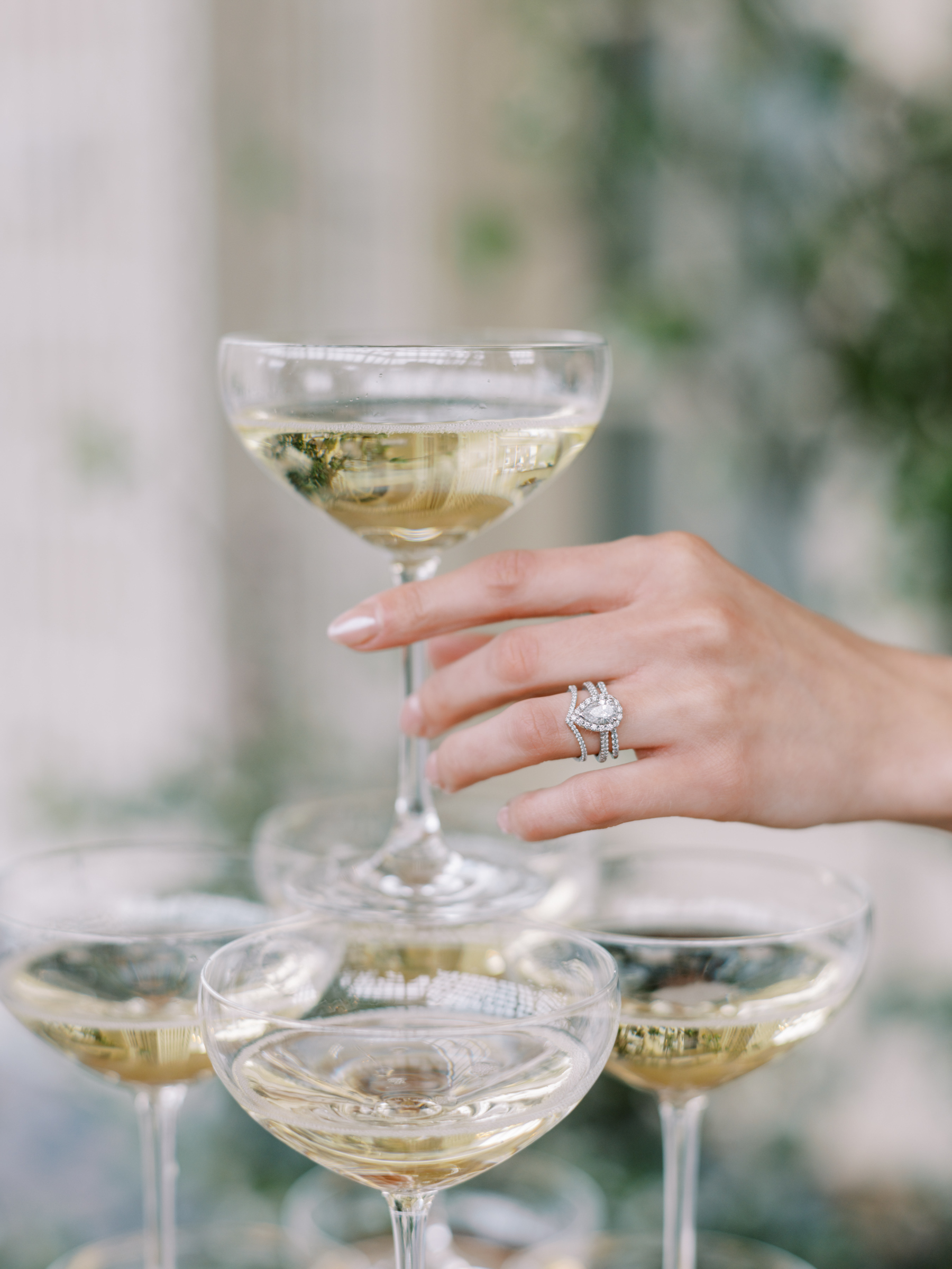 A hand with a diamond ring holds a coupe glass of sparkling wine above a stack of similar glasses—perfect inspiration for any wedding pro aiming to elevate their wedding business.