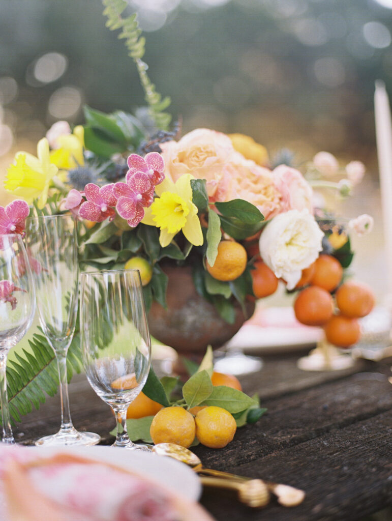 A floral centerpiece with yellow, pink, and white flowers, green leaves, and oranges sits on a wooden table set with glassware and plates—perfect inspiration for those looking to find your niche in the wedding industry.