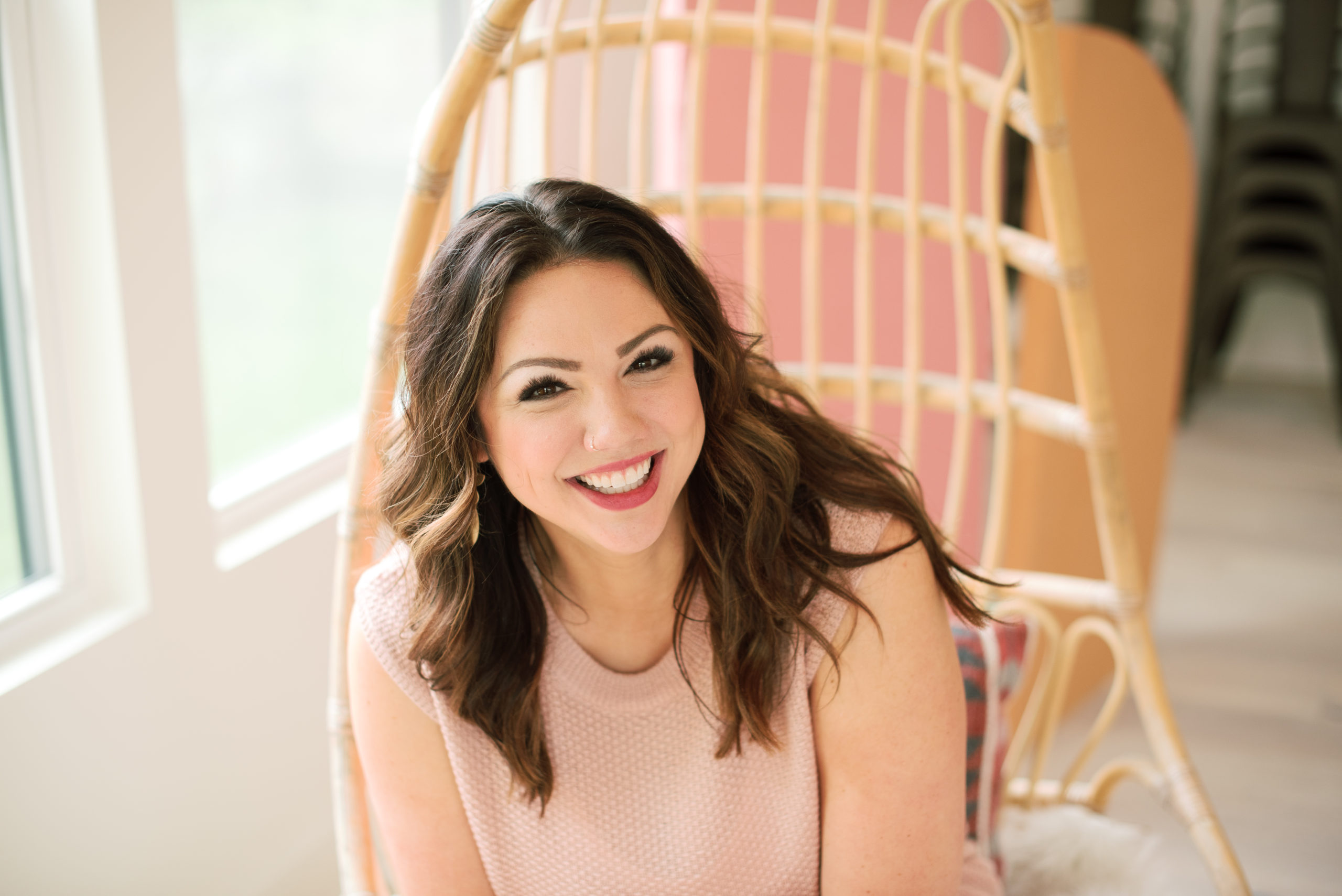 A woman with long brown hair sits on a wicker chair indoors, smiling toward the camera. She wears a light pink sleeveless top, looking relaxed and free from money stress often found in the wedding industry.
