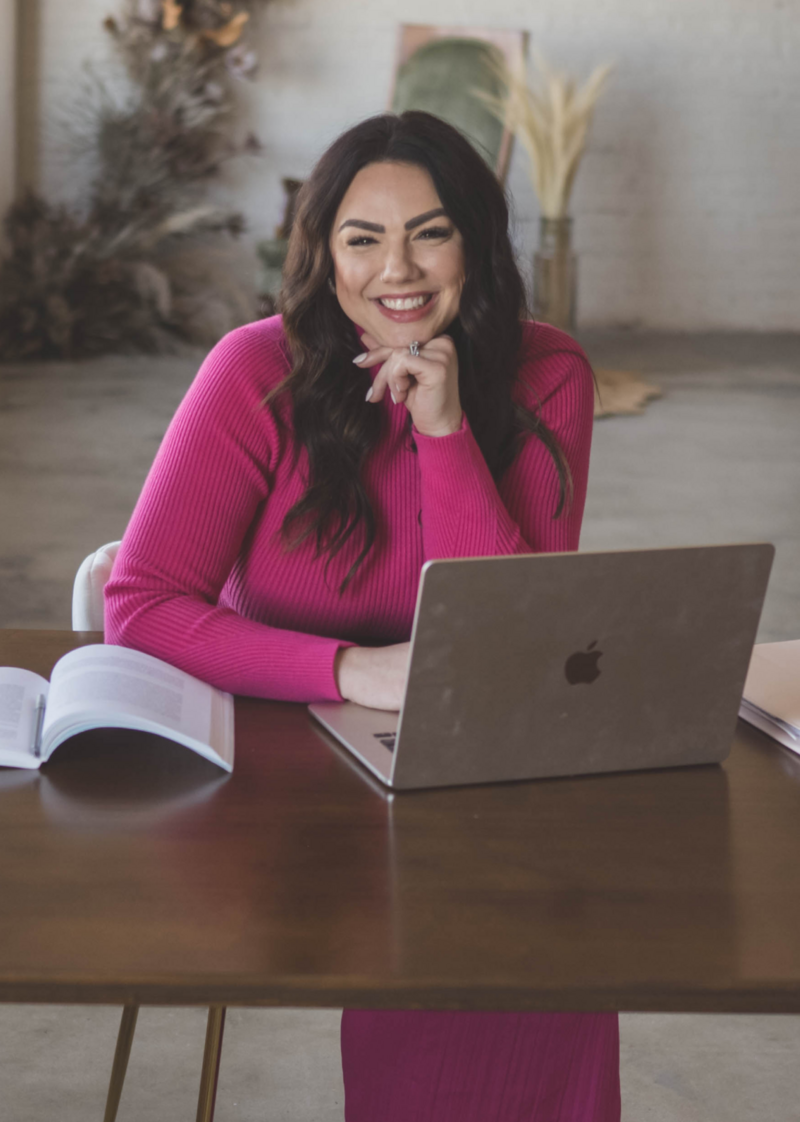 Woman in a bright pink outfit sits at a desk with an open laptop and notebook, smiling and resting her chin on her hand, ready to tackle wedding bookings in the busy wedding industry.
