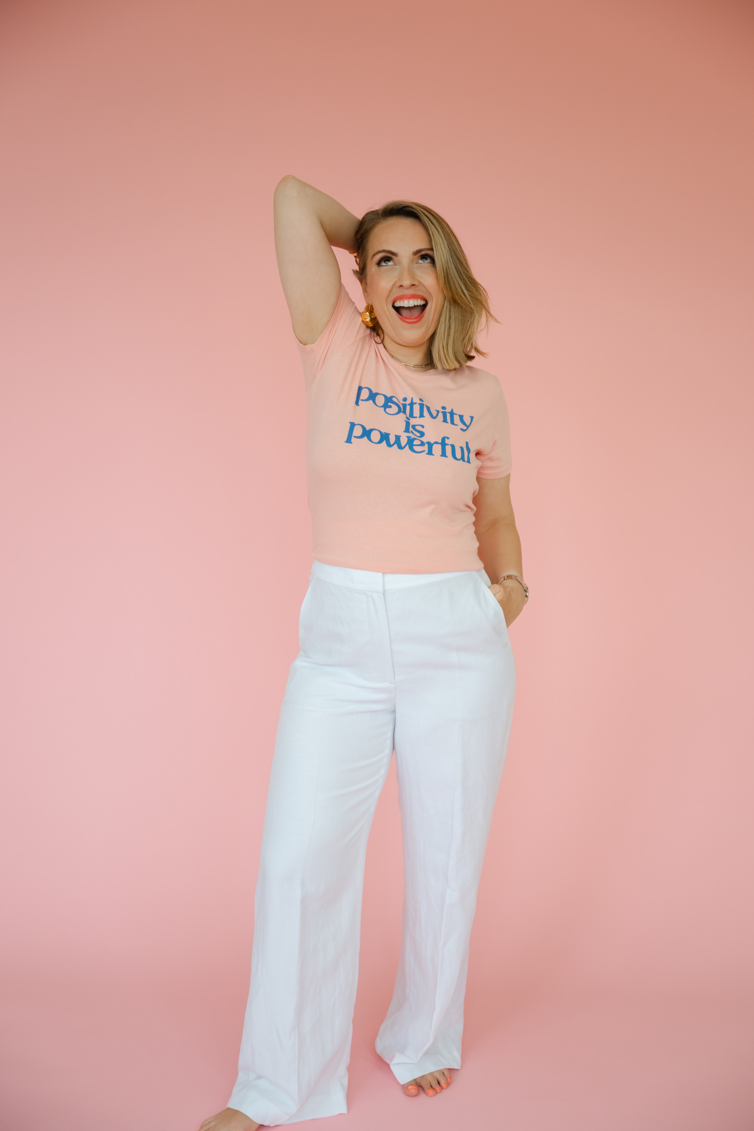 A woman poses barefoot against a pink background, wearing white pants and a peach shirt with the text "Positivity is powerful," embracing the confident spirit of high-achieving wedding pros.