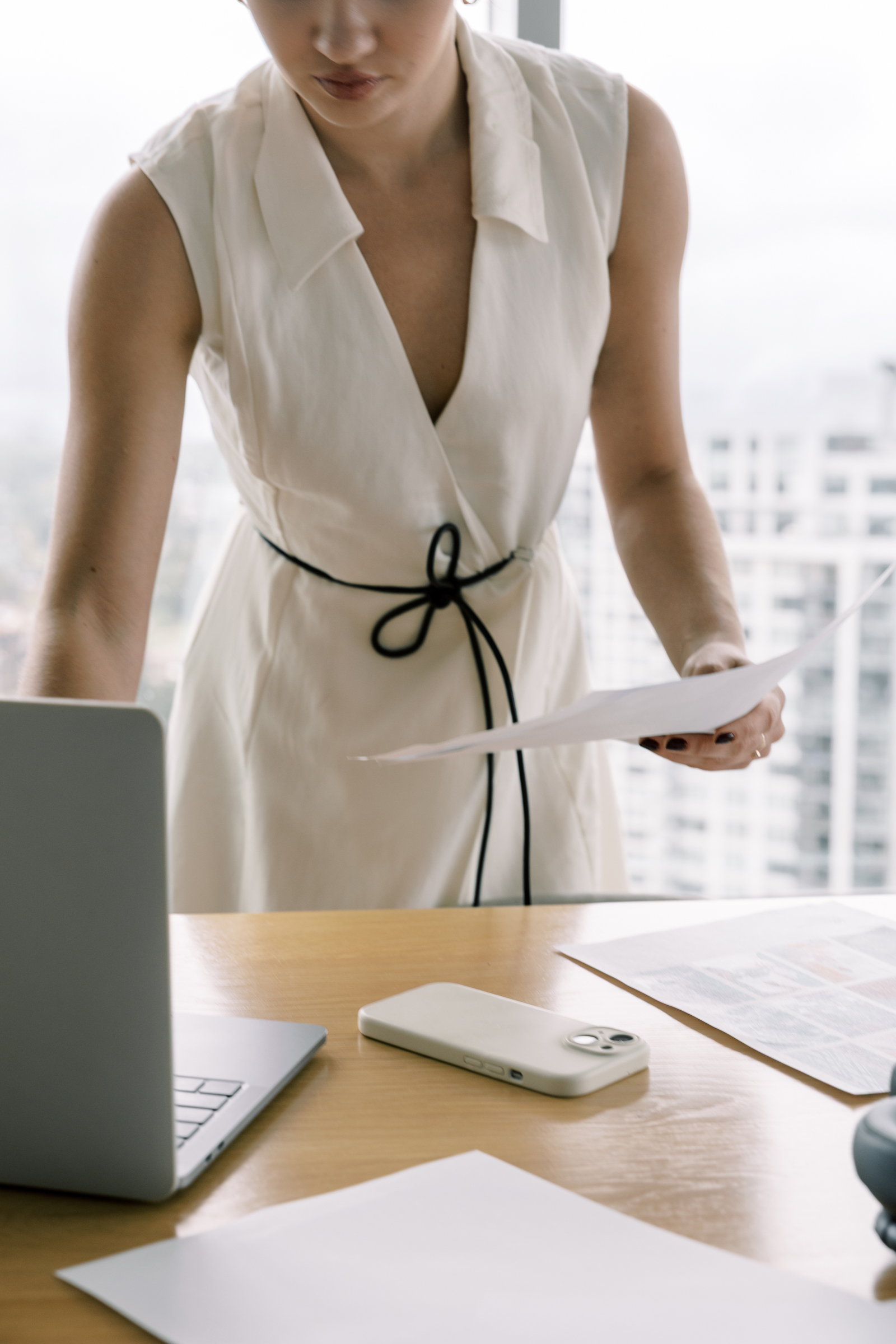 A woman in a white dress stands at a desk, holding papers and working on her laptop, with documents and a smartphone nearby—planning her growth strategy for wedding pros.