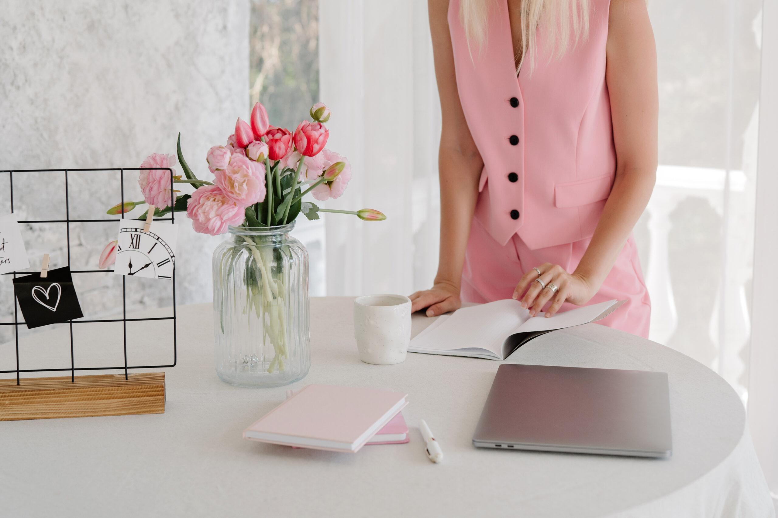 A person in a pink outfit stands at a round table with a laptop, mug, notebook, pen, vase of pink flowers, and a wire photo grid—perfectly capturing the start of a creative wedding planning 2026 journey.