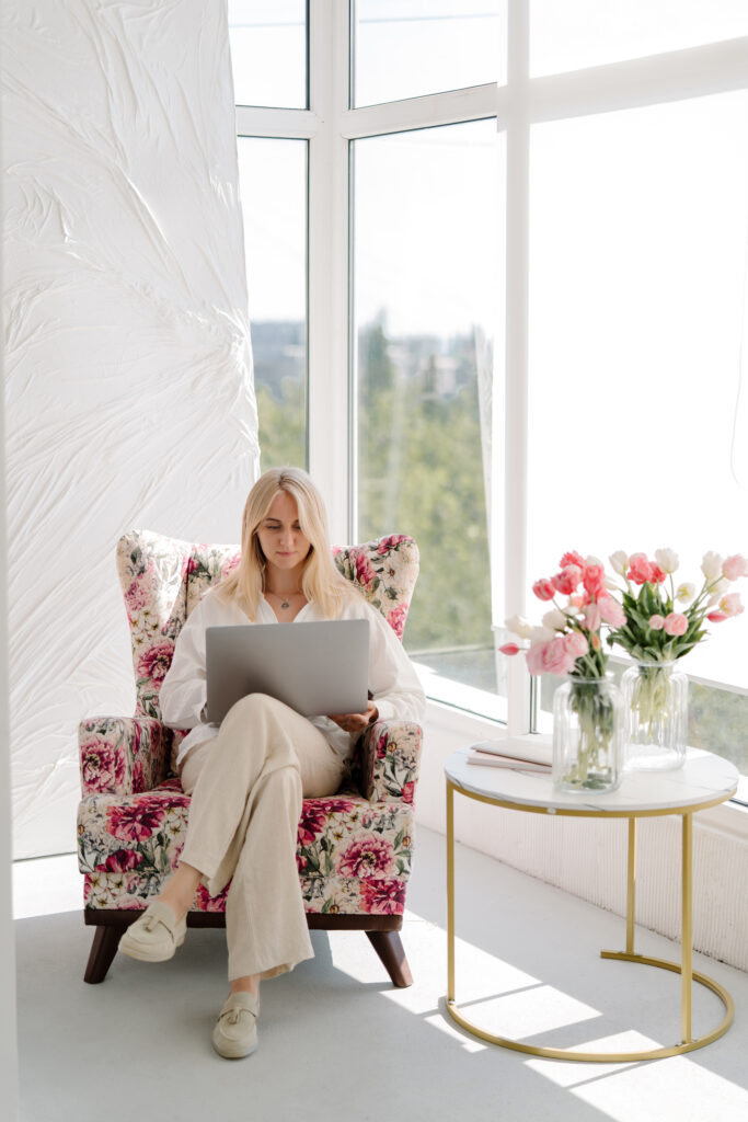 A woman sits in a floral armchair using a laptop&mdash;perhaps exploring wedding planning career opportunities&mdash;next to a round table with vases of pink and white flowers in a bright room with large windows.