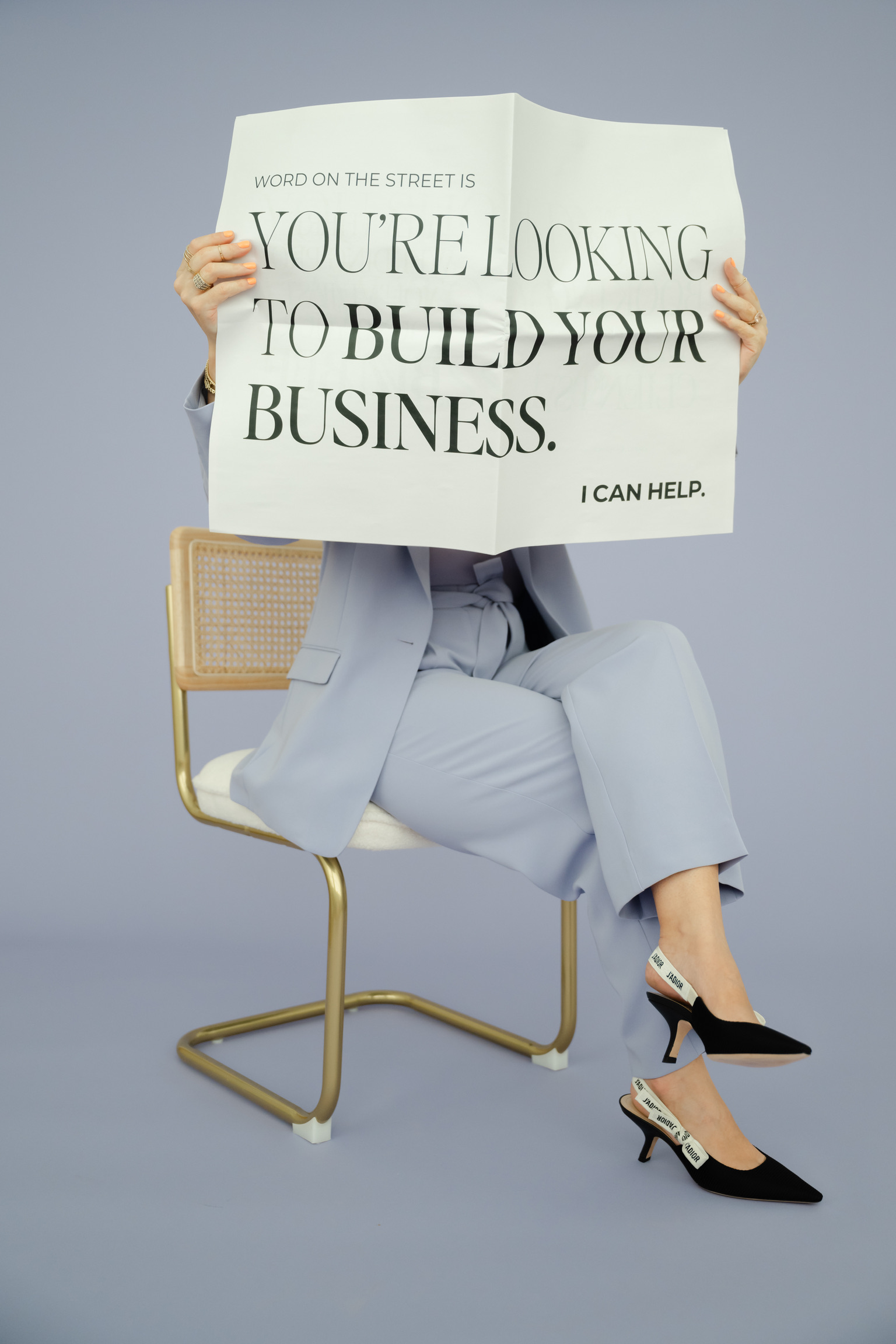 Person in a light blue suit sits on a chair holding a newspaper that reads, "YOU’RE LOOKING TO BUILD YOUR BUSINESS. I CAN HELP."—perfect inspiration for aspiring full-service wedding planners or anyone building their wedding planner checklist.