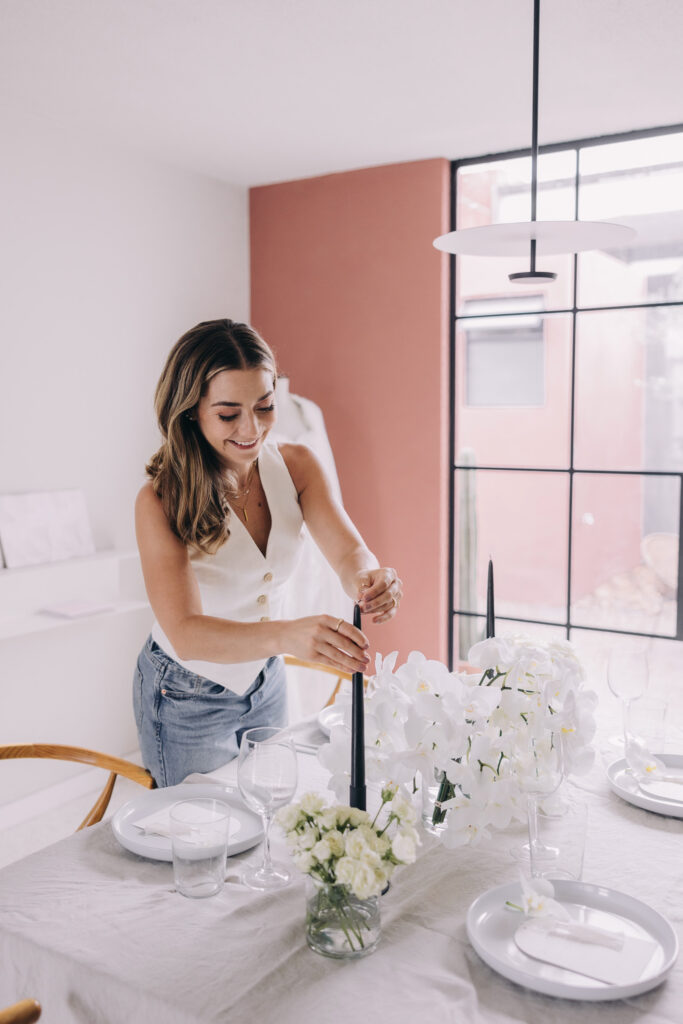 A woman sets a black candle into a candlestick on a dining table decorated with white flowers&mdash;an elegant touch that inspires those learning how to become a certified wedding planner in a modern, bright room.