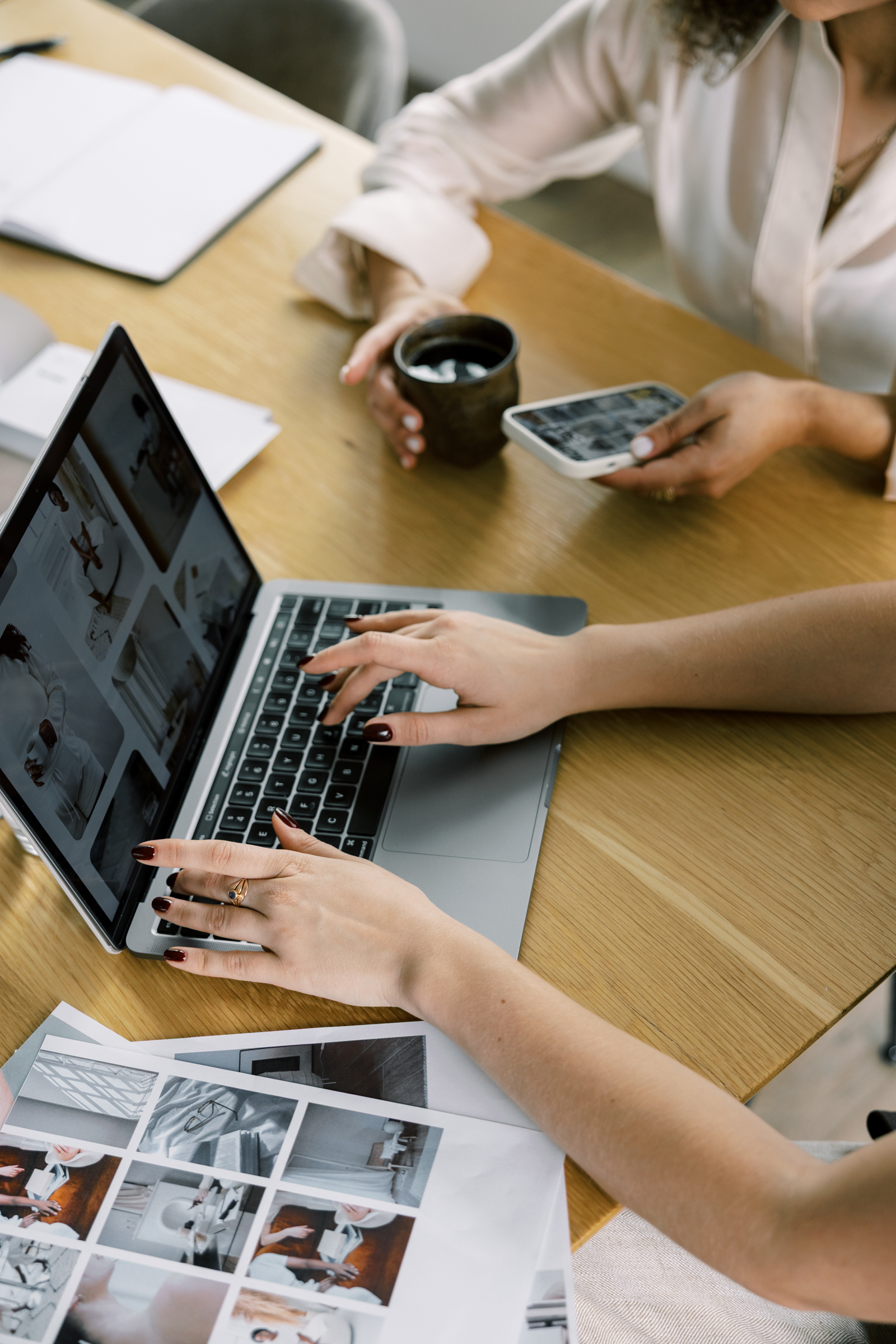 Two people work at a desk with a laptop displaying photo thumbnails, a smartphone, printed images, and coffee—collaborating on goal setting for wedding pros.