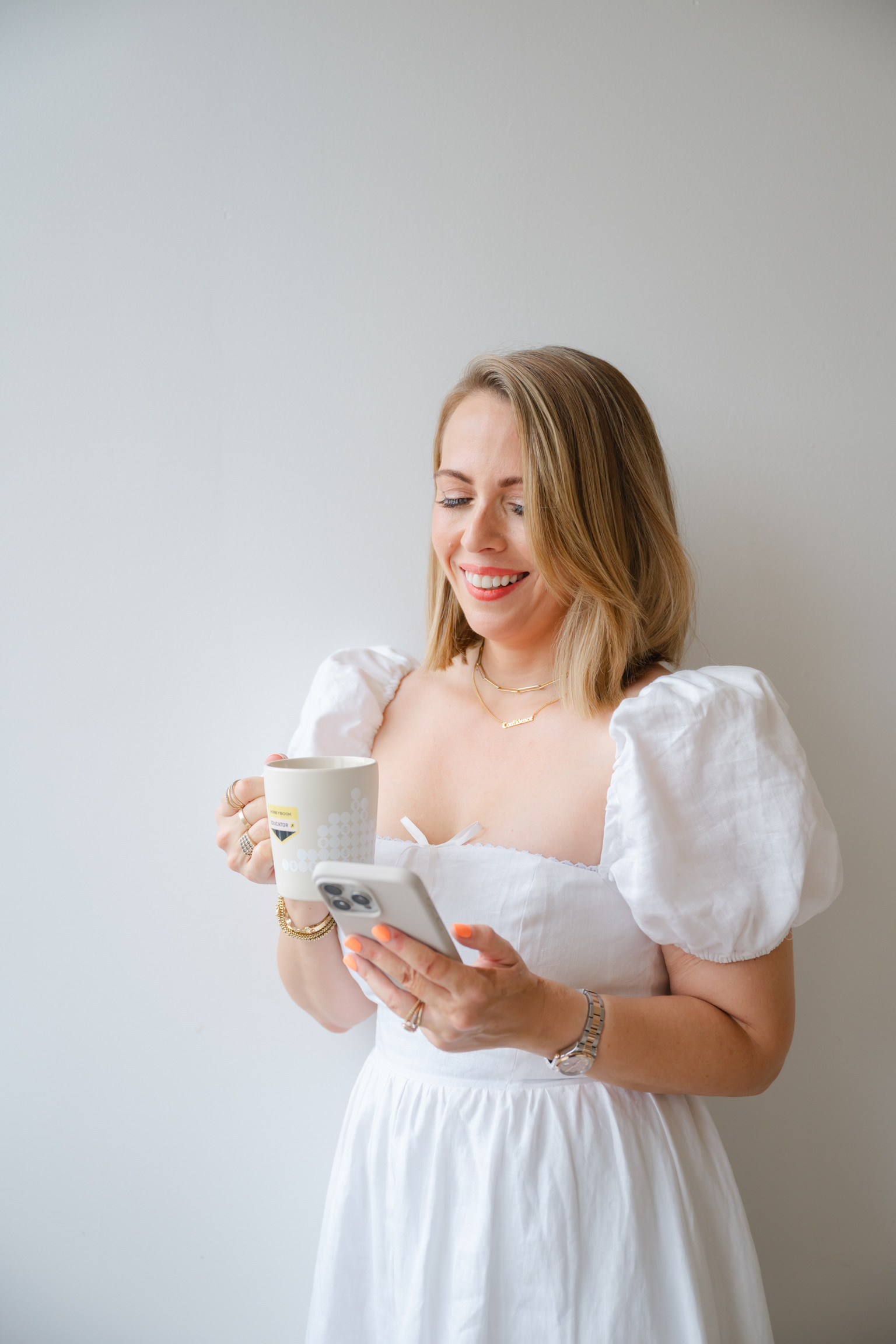 A woman in a white dress stands against a white wall, smiling while holding a mug and her smartphone—ready to share her latest wedding business advice.
