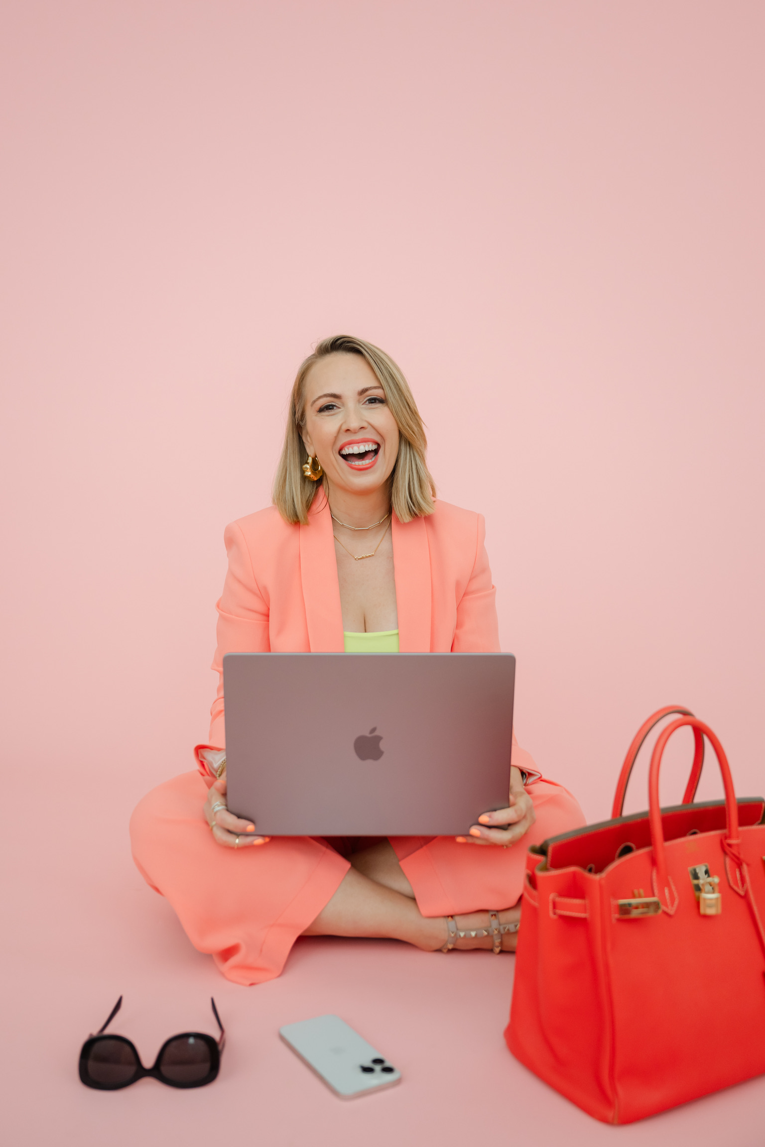 Woman in a bright coral suit sits cross-legged on the floor with a laptop, large matching handbag, sunglasses, and smartphone on a pink background—smiling broadly as if planning ideas for luxury wedding couples.