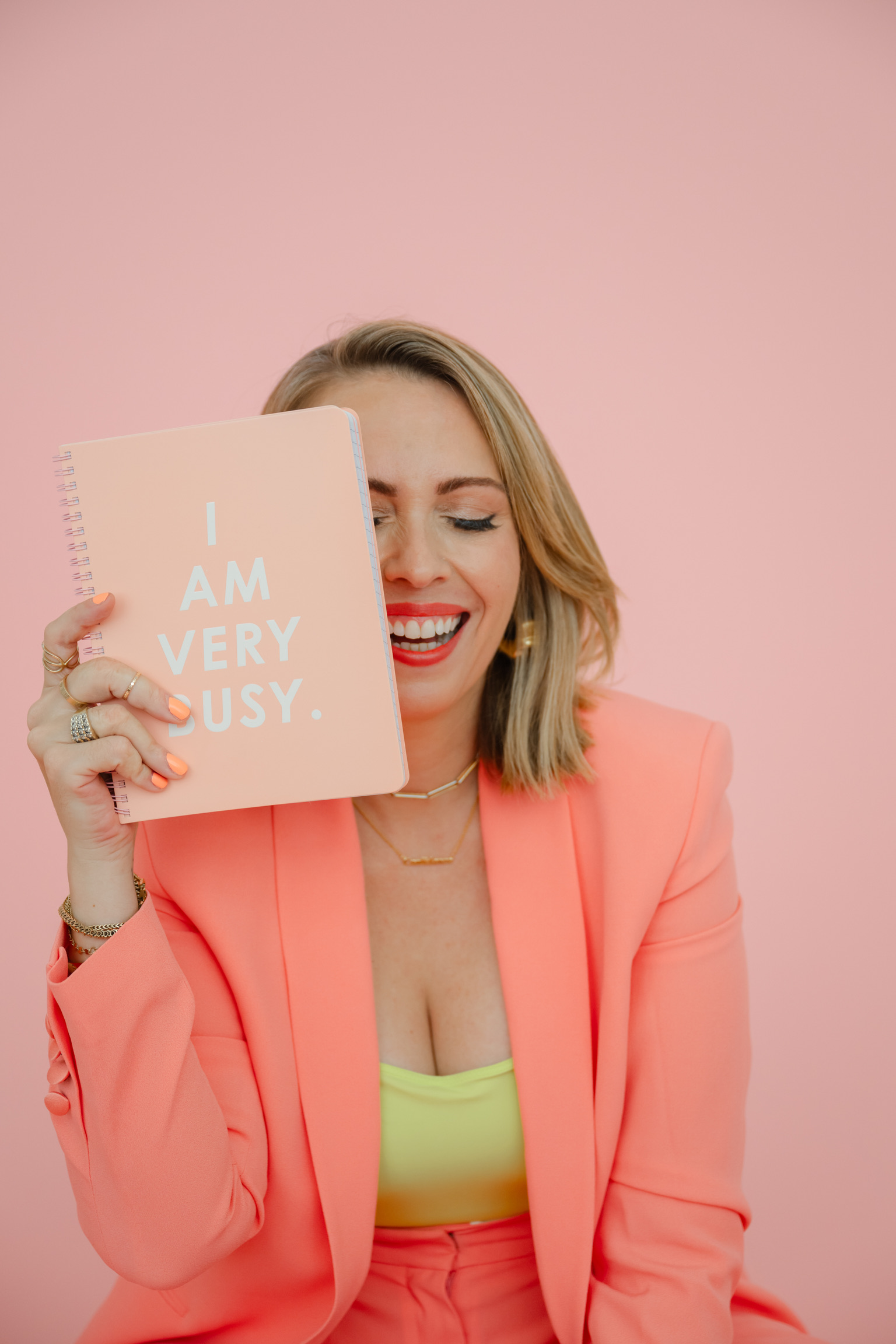 A woman in a coral suit holds a notebook labeled "I AM VERY BUSY" in front of her face, smiling with eyes closed, against a pink background—channeling effortless confidence and ready to share productivity hacks for wedding pros.