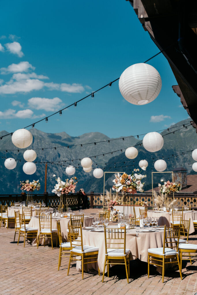 Outdoor event setup with round tables, gold chairs, floral centerpieces, and white paper lanterns hanging above&mdash;an ideal scene for those pursuing wedding planner education&mdash;set against mountains beneath a blue sky.