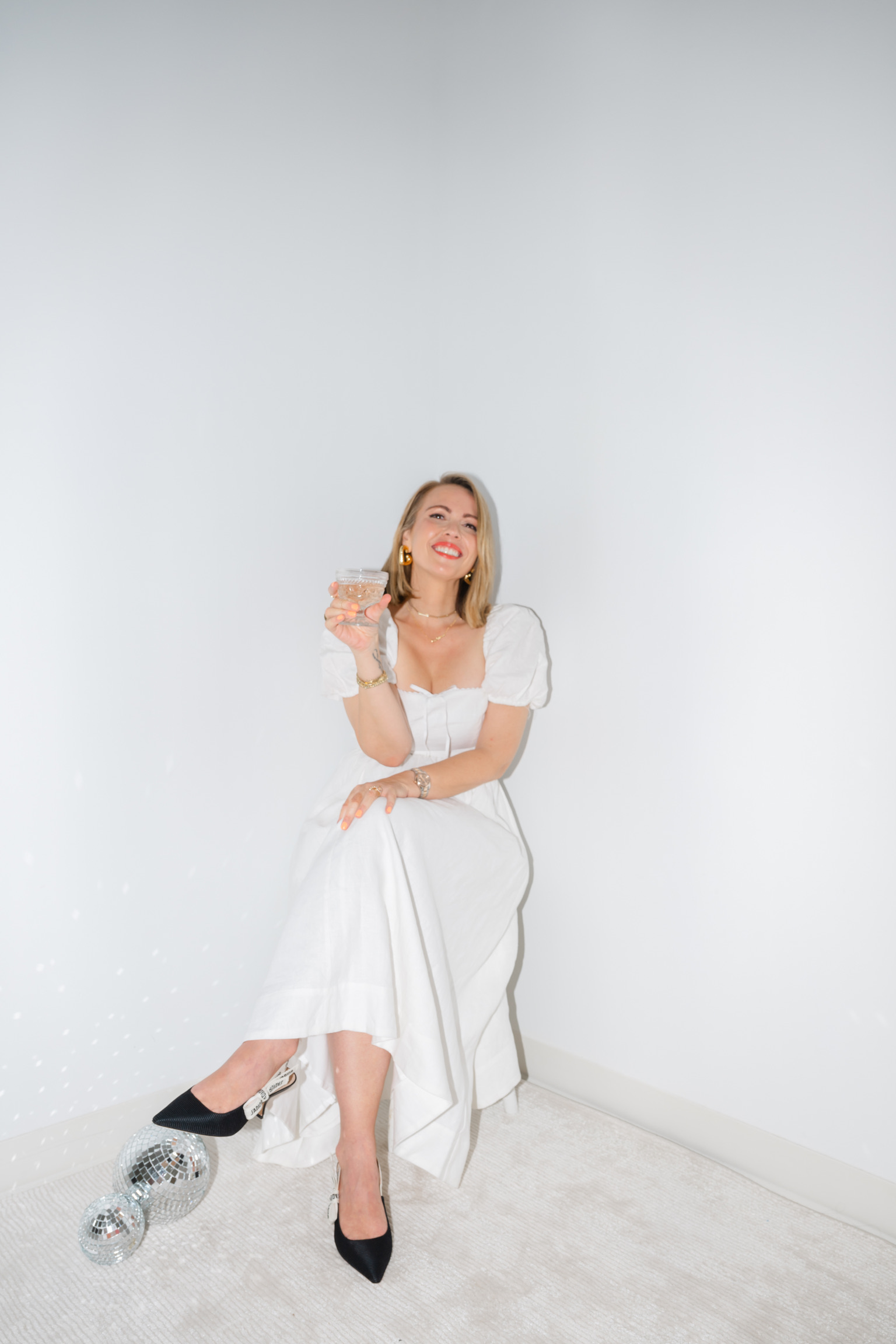 Woman in a white dress sits on a stool holding a drink, smiling like a wedding pro, with a disco ball on the floor against a plain white background—ready to plan your big day, whether your budget is $100K to $250K or you have just 12 months to go.