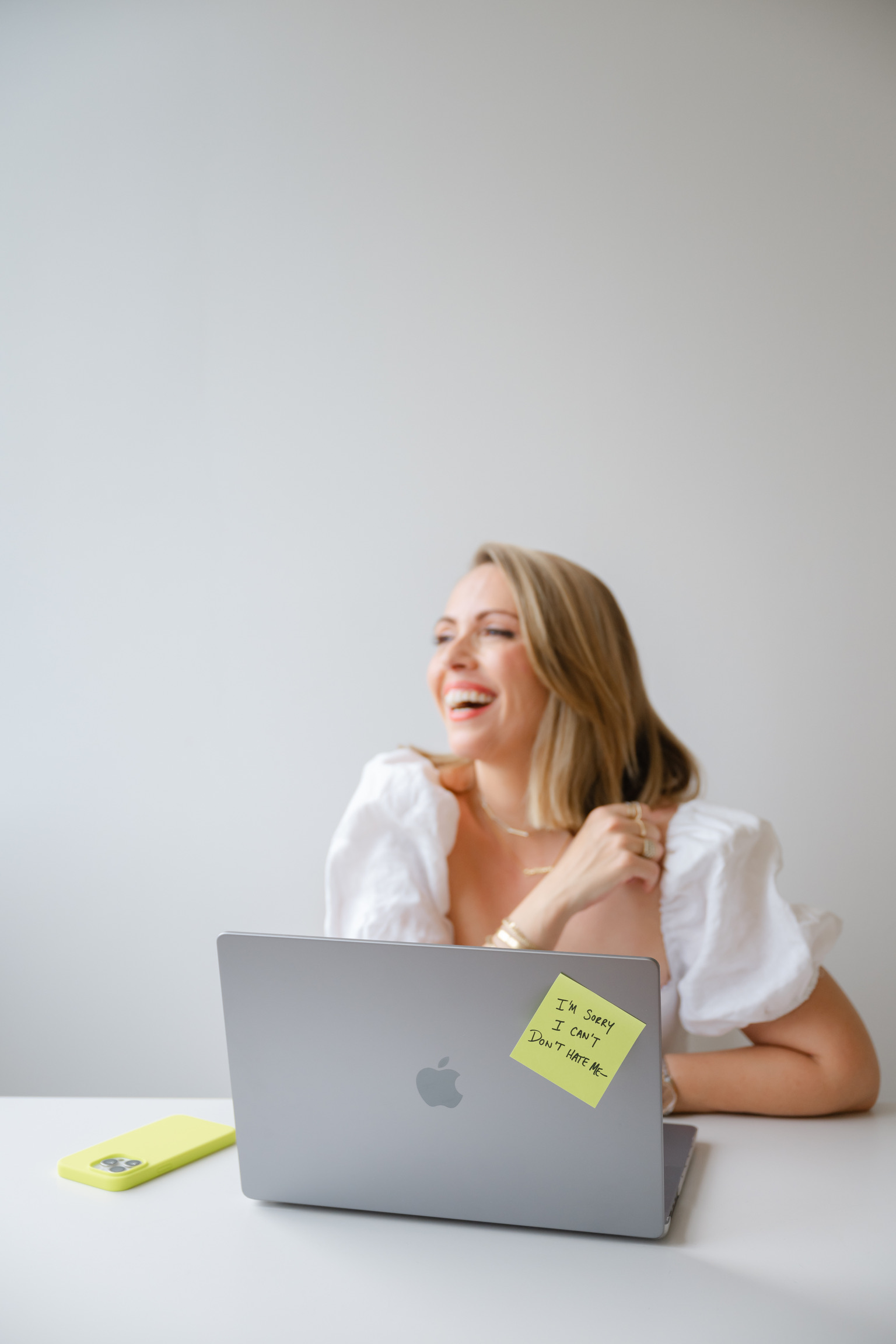 A woman sits at a desk with a laptop, smiling. A yellow phone and a sticky note reading "I swear I can't. Don't hate me." hint at Nervous System Dysregulation in the Wedding Industry.