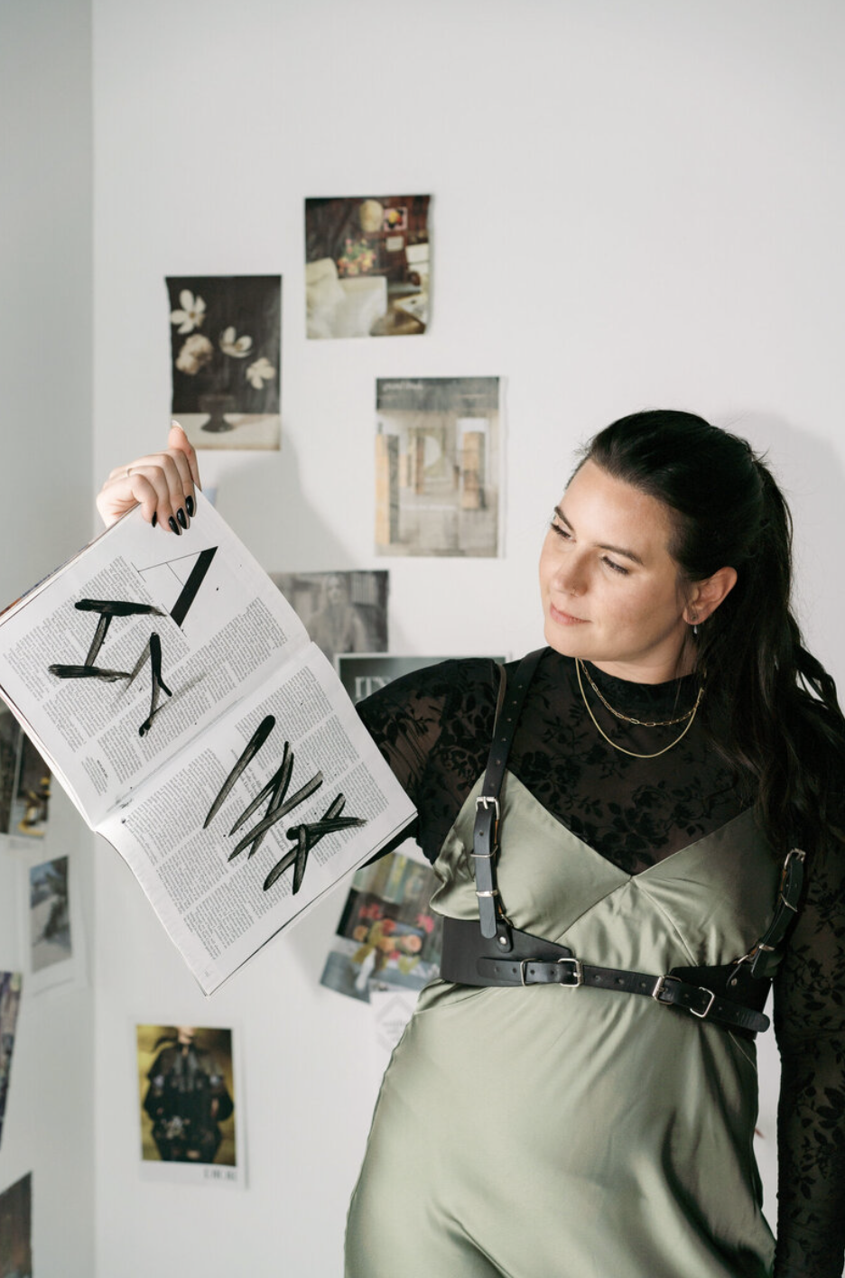 A woman in a green dress and black harness holds up an open magazine with black paint marks, standing in front of a wall with various photos and artwork.