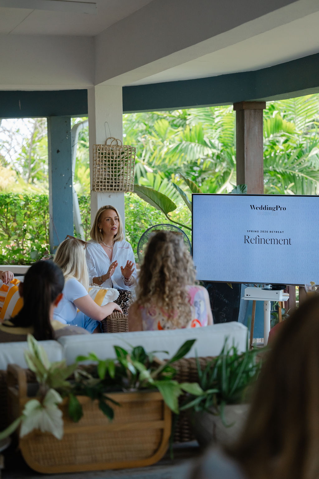 A woman gives a presentation to a seated group, with a screen displaying "WeddingPro," "Refinement," and tips for building a Six-Figure Wedding Business in a bright indoor-outdoor setting filled with plants.