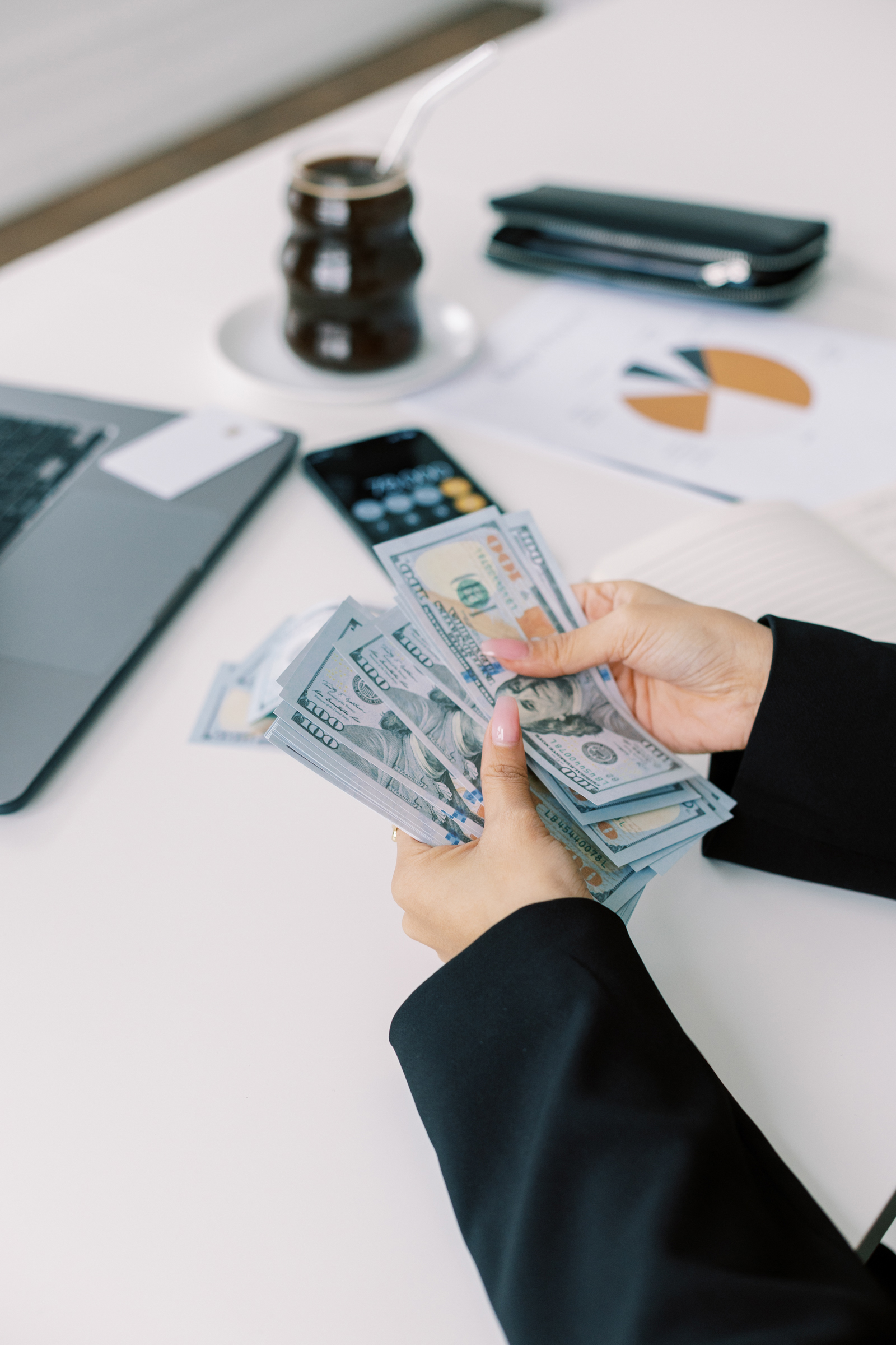 Person counting several $100 bills at a desk with a laptop, documents, and phone—measuring ROI on wedding business decisions as they review their earnings.
