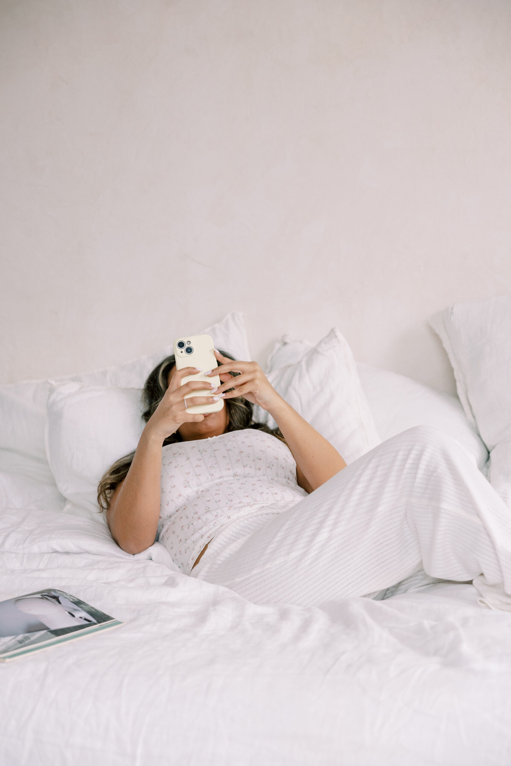 A person lies on a white bed, holding a smartphone and looking at the screen—perhaps checking messages about wedding client management. A magazine is open on the bed nearby.