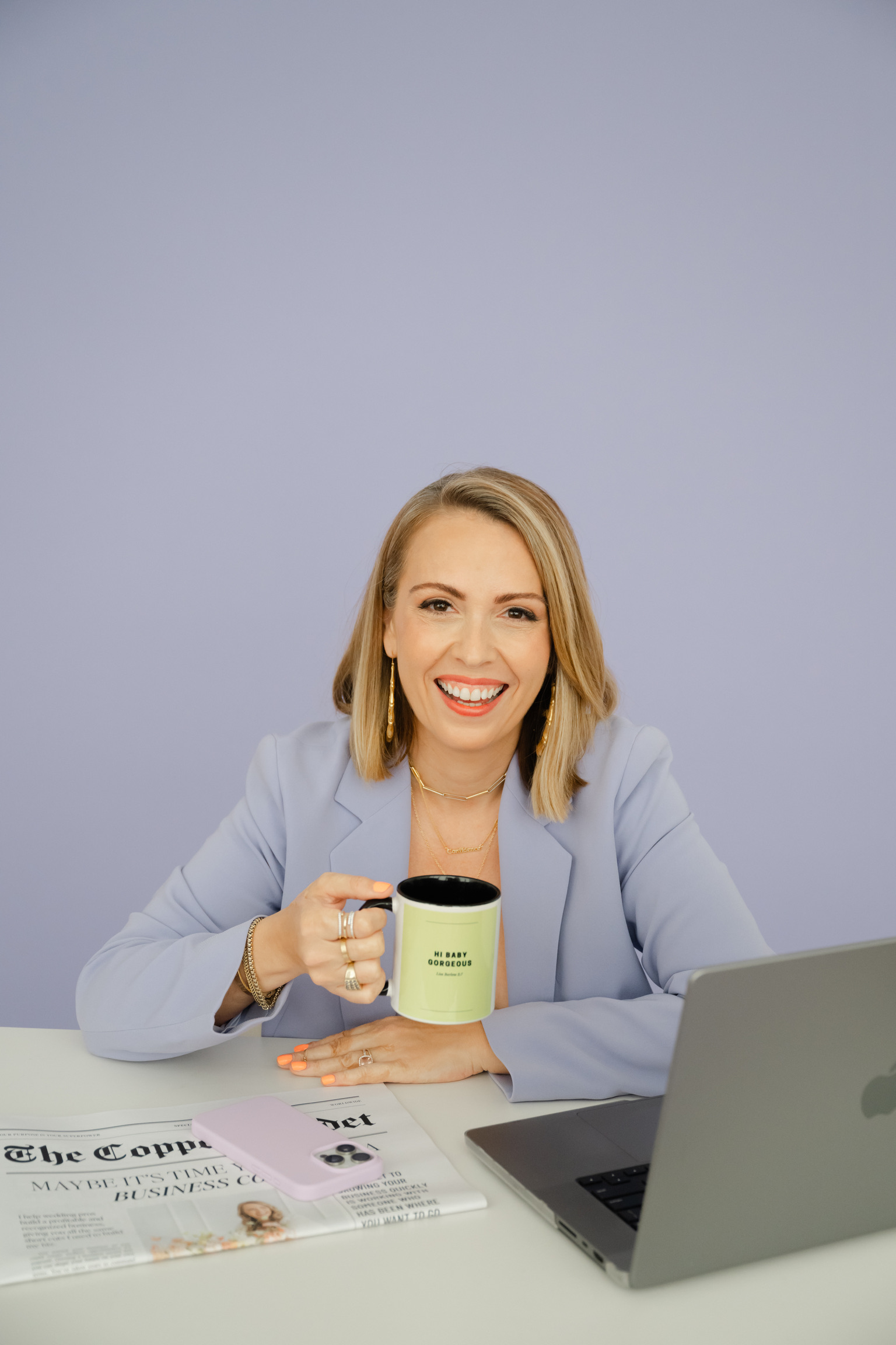 Woman in a lavender blazer sits at a desk with a laptop, holding a coffee mug and smiling. Newspapers are spread out on the desk in front of her as she prepares for launching a wedding planning software.