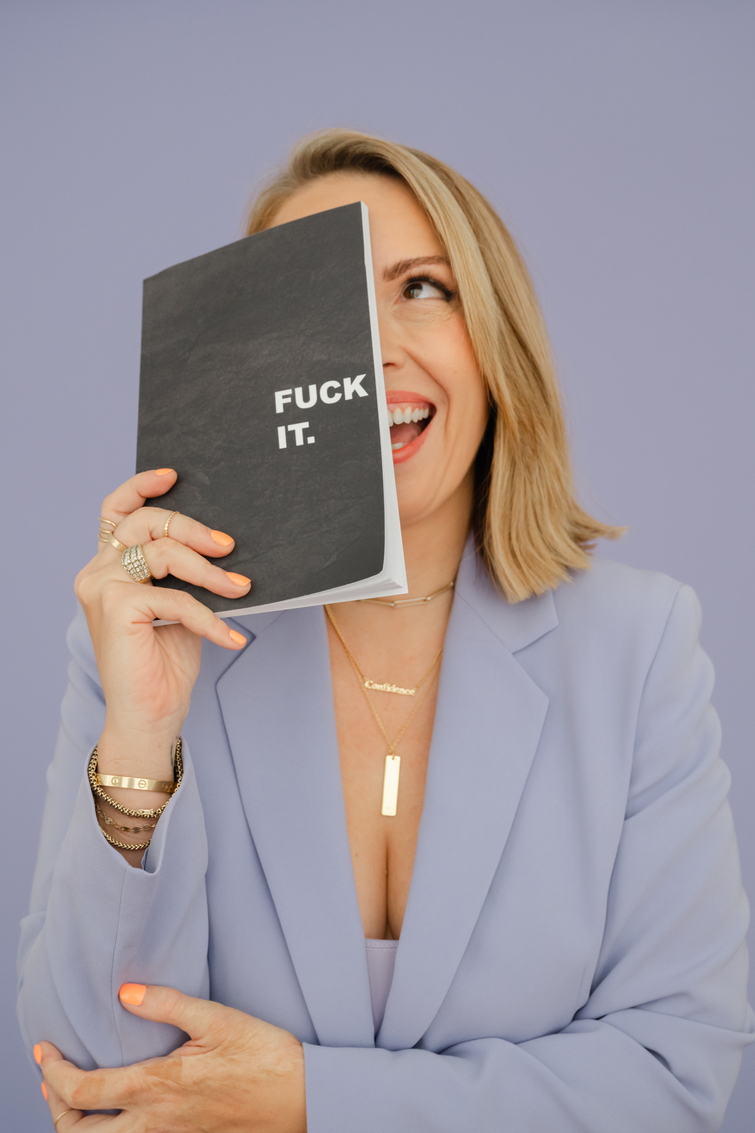 A woman in a light purple blazer holds a black notebook that says “FUCK IT” in white letters, partially covering her face against a plain lavender background.