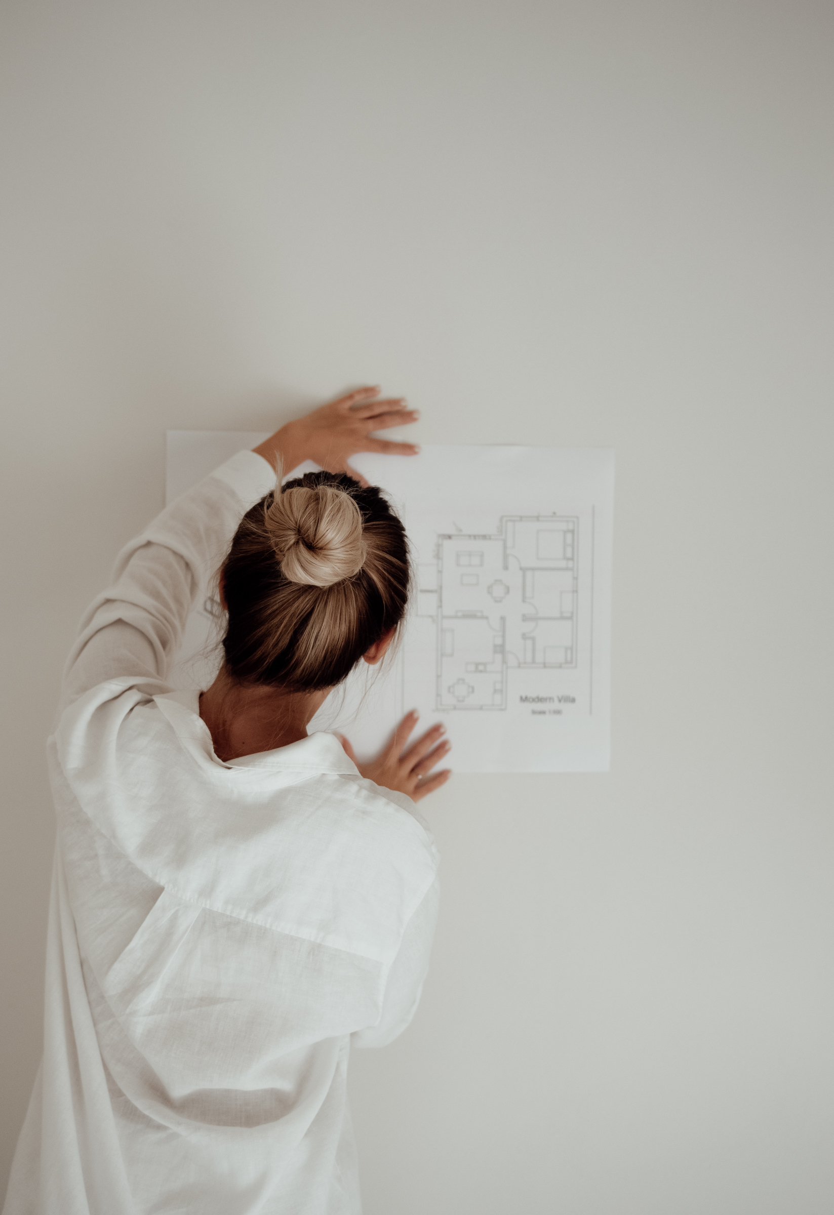 A person with hair in a bun, seen from behind, pins a floor plan labeled “Modern Villa” to a plain white wall—an inspiring space for those looking to Price Your Wedding Services with elegance and professionalism.