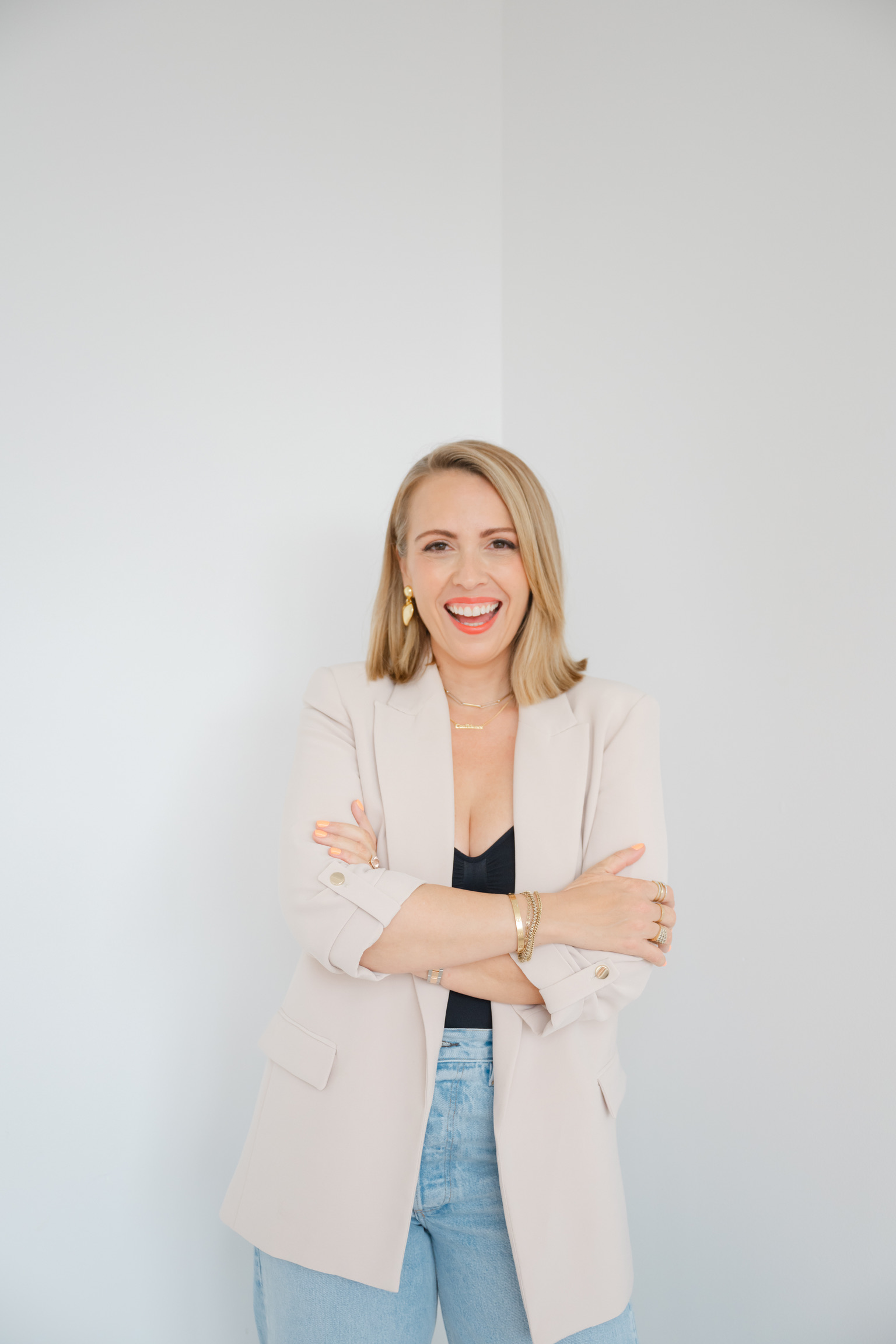 A smiling woman with blonde hair, wearing a beige blazer, black top, and blue jeans with her arms crossed, ready to make strategic business decisions.